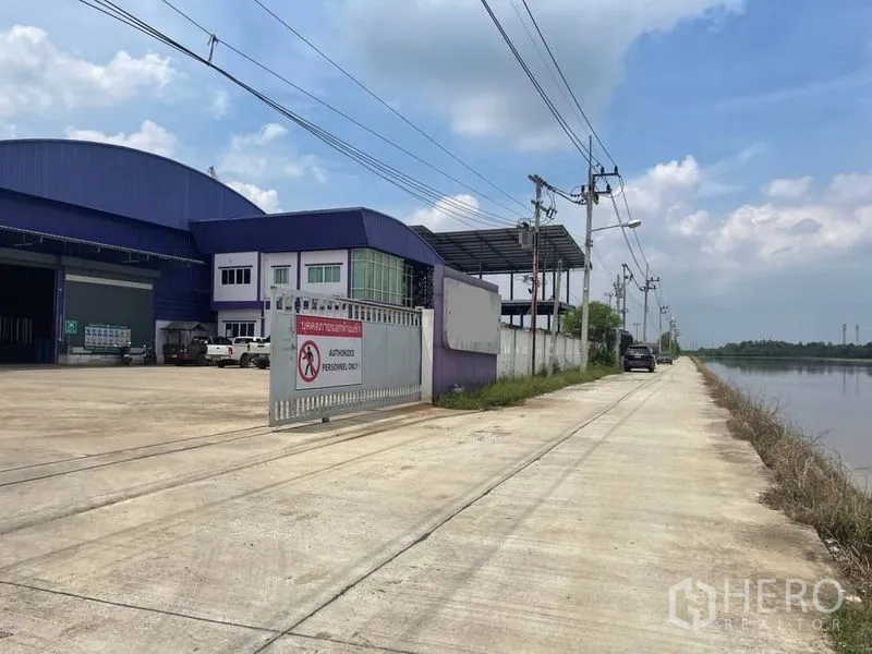 Warehouse for sale in Lam Luk Ka, Pathum Thani - Gated warehouse entrance on a canal-side road with perimeter wall, security sign and overhead power lines.