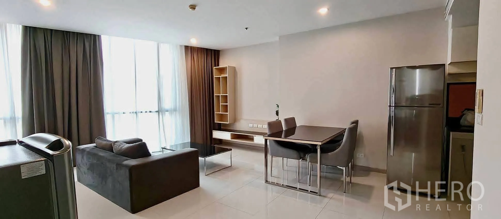 Condominium for rent in Watthana, Bangkok - Bright lounge with floor-to-ceiling windows, dark curtains, and adjacent dining area beside a refrigerator.