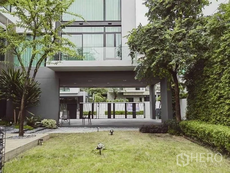 บ้าน ให้เช่า - สนามหญ้านำสู่ที่จอดรถมีหลังคา ใต้ชั้นบนยื่นลอย รายล้อมด้วยแนวรั้วต้นไม้.