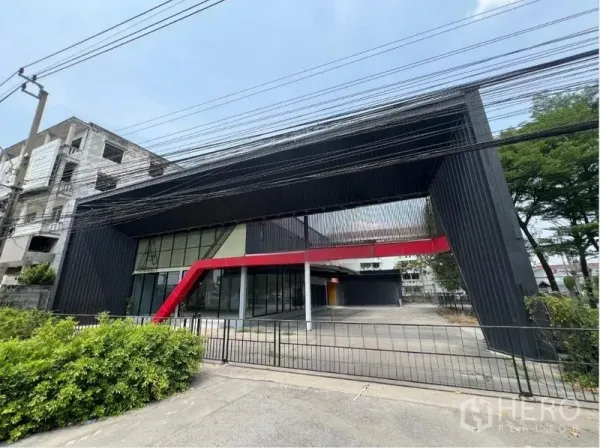 零售空间 出租在Saphan Sung, Bangkok - 现代黑色立面零售建筑，红色装饰梁与整面玻璃展厅，前方有围栏庭院。