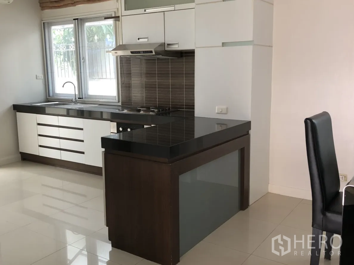 บ้าน ให้เช่าหรือขายในLat Phrao, Bangkok - ครัวรูปตัวแอล เคาน์เตอร์หินแกรนิตสีดำ ตู้สีขาว และผนังกระเบื้องสีน้ำตาล