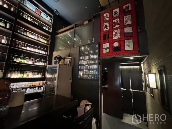 店屋 出租在Watthana, Bangkok - 吧台后侧特写，饮料陈列架、冰箱与通往夹层的楼梯，红色装饰墙。