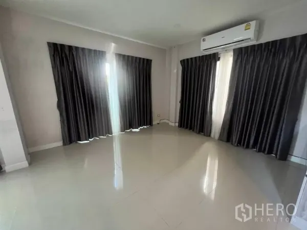 Detached House for rent in Samut Prakan - Bright empty living room with glossy tile floors, dark curtains and a wall-mounted air conditioner.