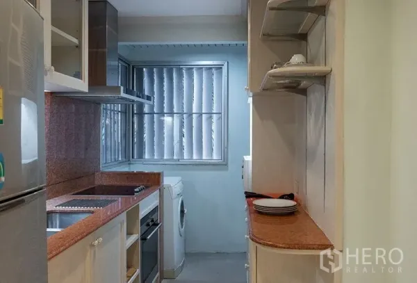 Apartment for rent in Bang Rak, Bangkok - Compact galley kitchen with granite countertops, electric hob, oven, range hood, shelves and a washing machine by a louvered window.