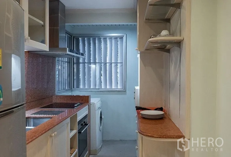 Apartment for rent in Bang Rak, Bangkok - Compact galley kitchen with granite countertops, electric hob, oven, range hood, shelves and a washing machine by a louvered window.