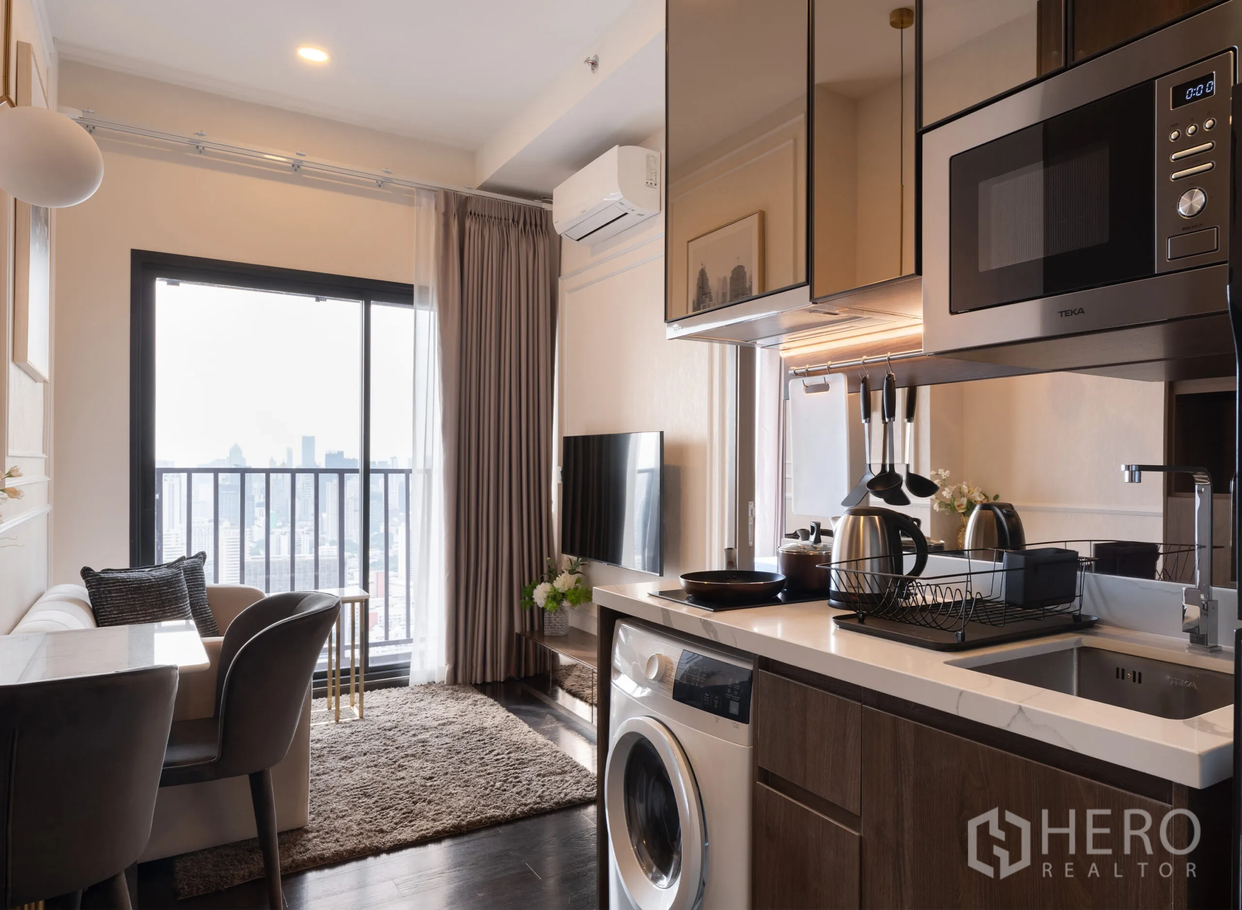 公寓 for rent in Watthana, Bangkok - 开放式客厨一体，带橱柜、微波炉、洗衣机、餐桌椅、沙发、电视与阳台城市景观。