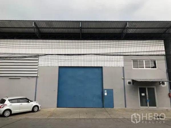 货栈 出租在Bang Kapi, Bangkok - 曼谷Bang Kapi仓库外观，配大型蓝色装卸卷闸门与带雨棚的玻璃办公室入口。