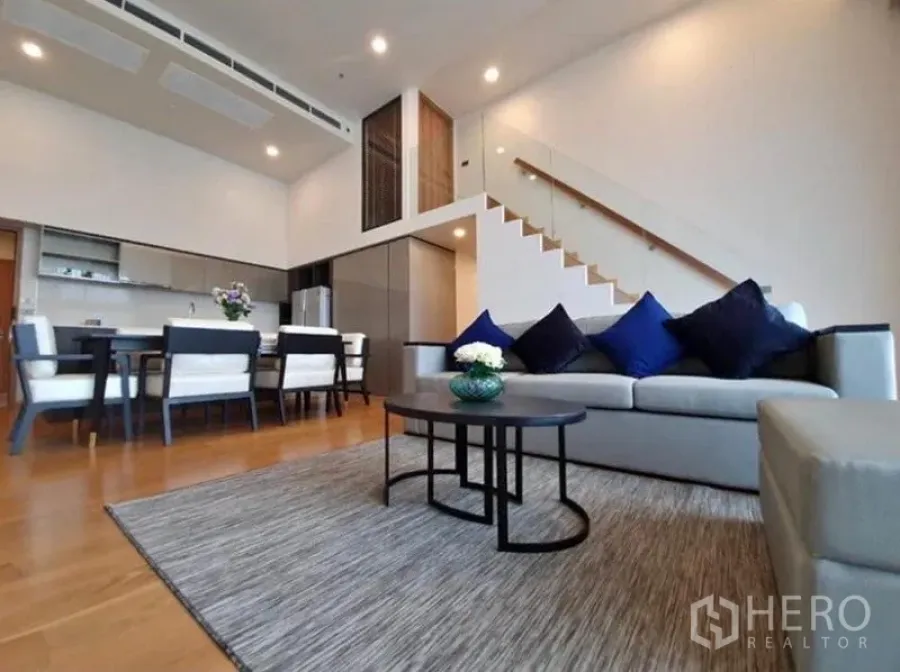 Condominium for rent in Watthana, Bangkok - Open-plan living and dining room with double-height ceiling, glass staircase, and modern kitchen in a Bangkok condo.