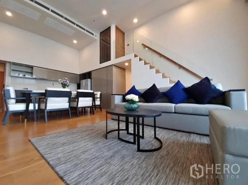 Condominium for rent in Watthana, Bangkok - Open-plan living and dining room with double-height ceiling, glass staircase, and modern kitchen in a Bangkok condo.