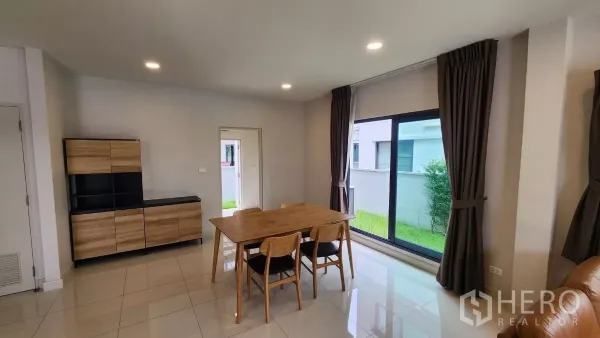 Detached House for rent in Bang Phli, Samut Prakan - Dining area with wooden table for four beside wide windows overlooking a green side yard.