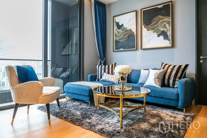 Condominium for rent or sale in Khlong Toei, Bangkok - Bright living room with blue sofa, gold glass coffee table, and floor-to-ceiling windows overlooking Bangkok.