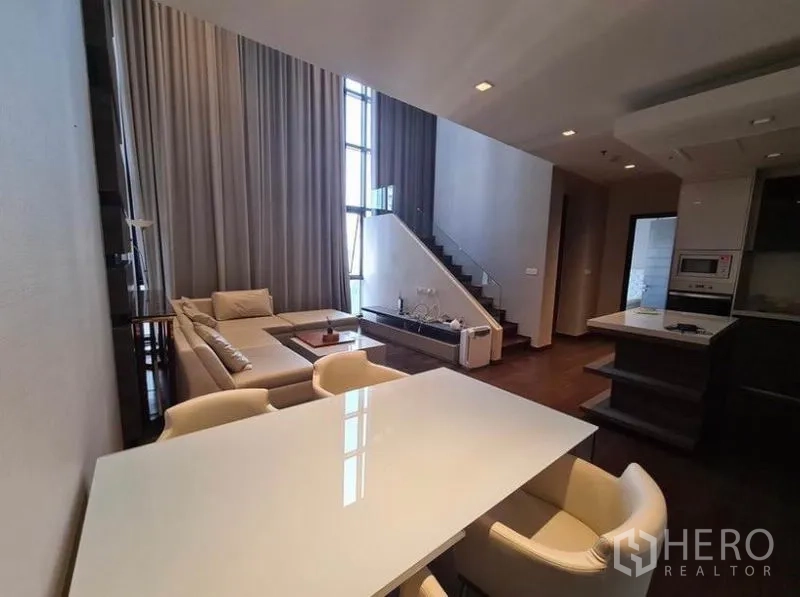 Condominium for rent or sale in Huai Khwang, Bangkok - Dining table facing lounge and staircase with high windows bringing natural light.