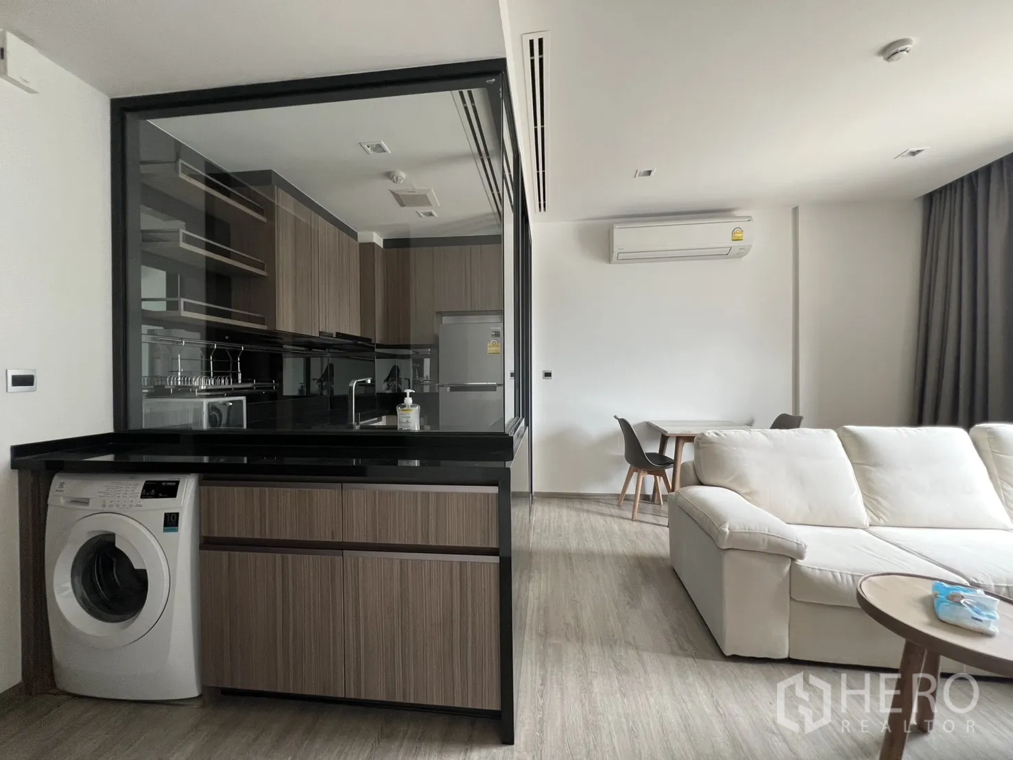 公寓 for rent in Watthana, Bangkok - 玻璃隔断后的现代厨房，黑色台面，面向白色沙发与小餐桌。