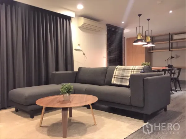 Condominium for rent or sale in Watthana, Bangkok - Cozy living room featuring a large gray L-shaped sofa, round coffee table, and floor-to-ceiling curtains.