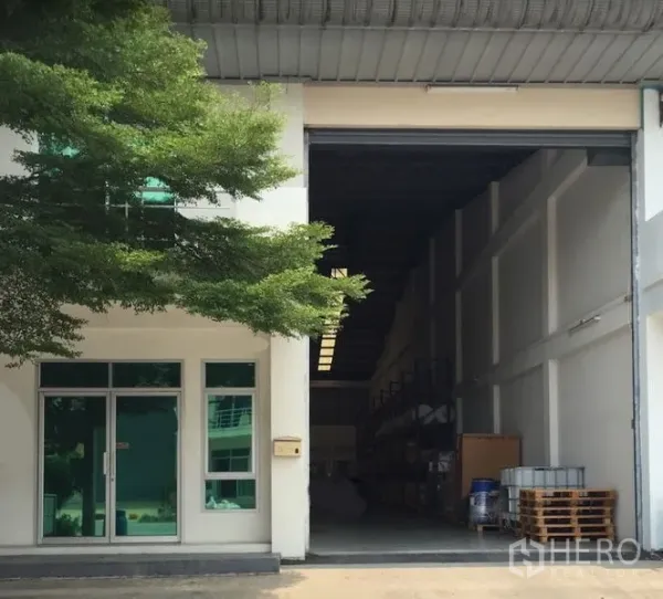 Factory for rent in Mueang Samut Prakan, Samut Prakan - Factory frontage with glass office, shade tree and large roller shutter opening to a high-ceiling warehouse.