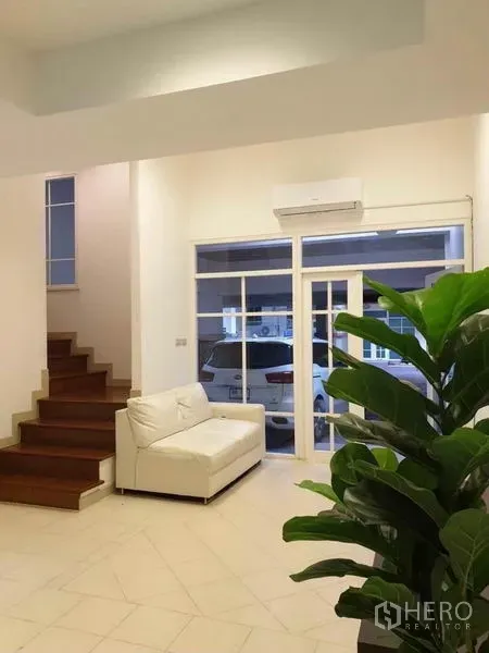 ทาวน์เฮาส์ ให้เช่าหรือขายในChatuchak, Bangkok - ห้องนั่งเล่นโปร่งสว่าง โซฟาสีขาว บันไดไม้ ต้นไม้ในบ้าน และประตูกระจกออกสู่ที่จอดรถในร่ม.