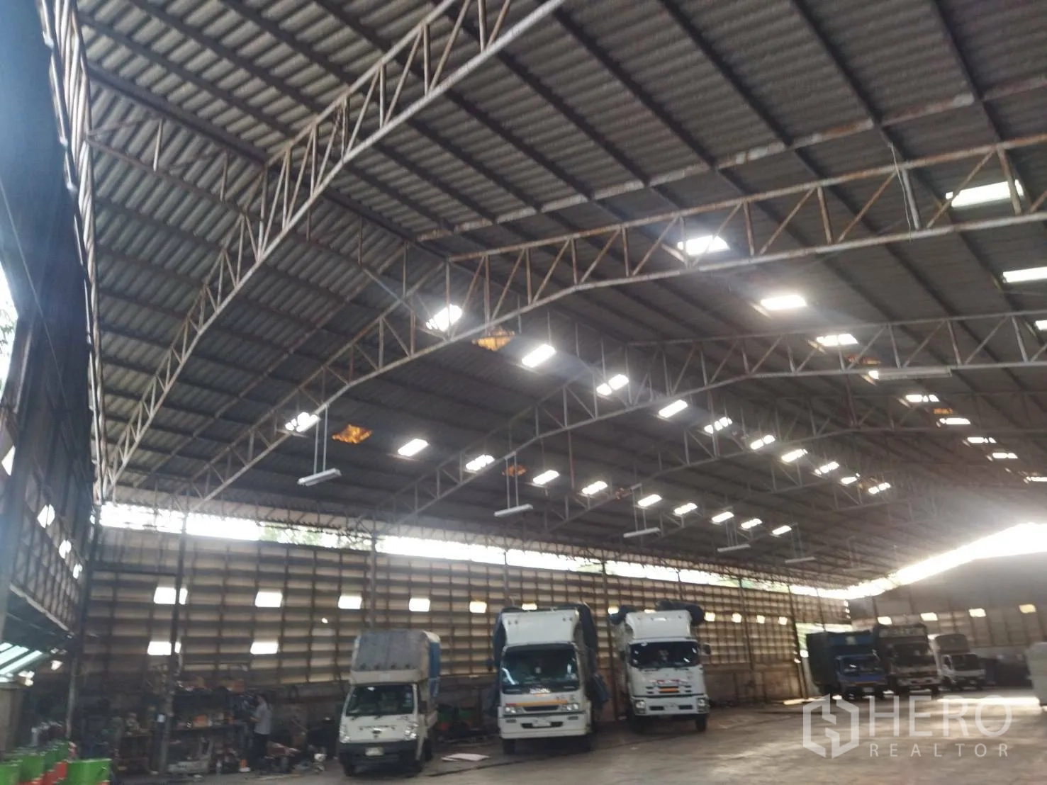 Warehouse for rent in Bang Khun Thian, Bangkok - Spacious warehouse interior with high steel truss roof, skylights, and trucks parked for loading.