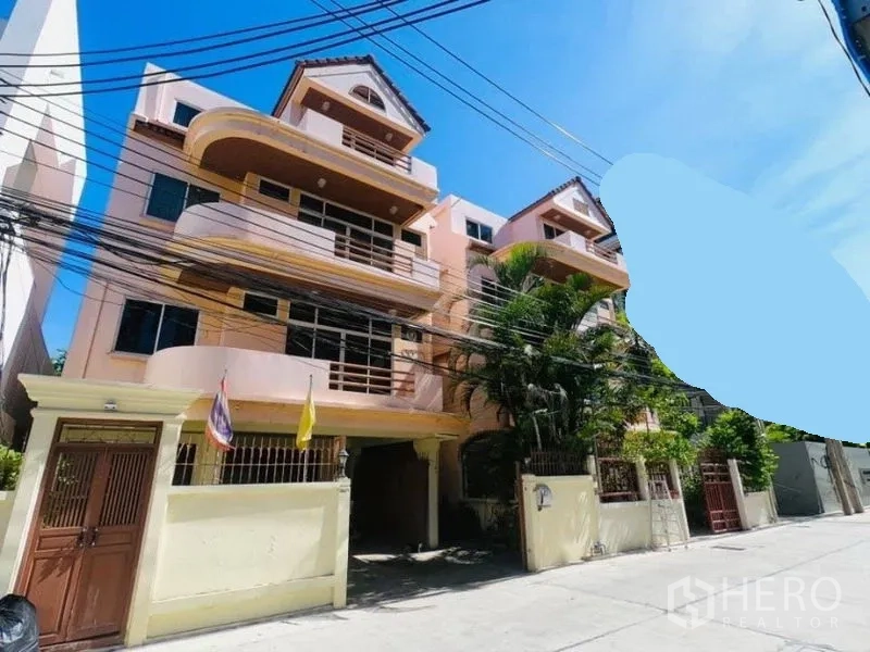 บ้าน ให้เช่าในWatthana, Bangkok - ด้านหน้าบ้านพร้อมระเบียงหลายชั้นและประตูรั้ว พร้อมต้นไม้ทรอปิคัล