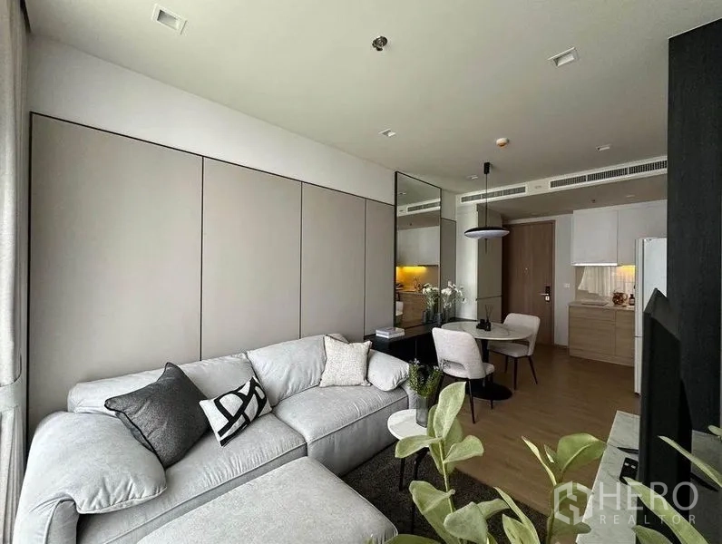 Condominium for rent in Watthana, Bangkok - Open-plan living and dining area with L-shaped sofa, mirror feature wall, compact kitchen and wood flooring.