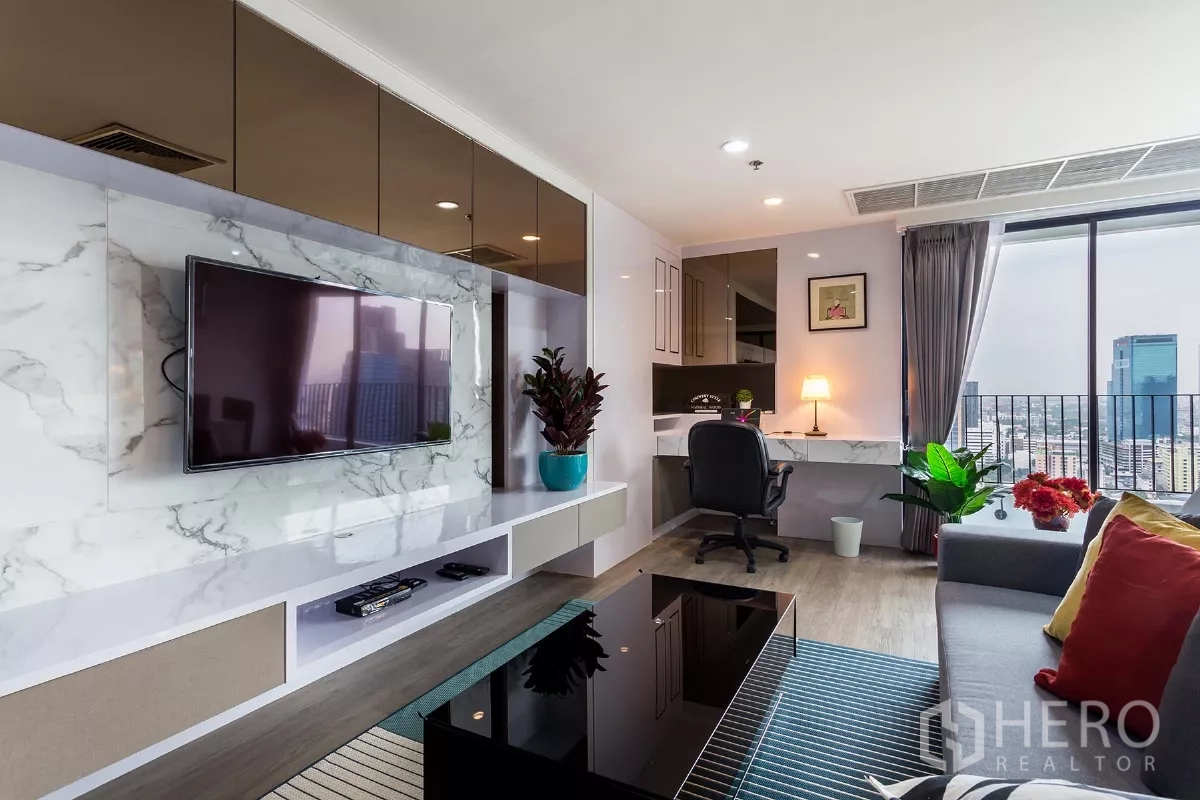 Condominium for rent or sale in Watthana, Bangkok - Living room with wall-mounted TV on a marble feature wall and a work desk by the window with city view.