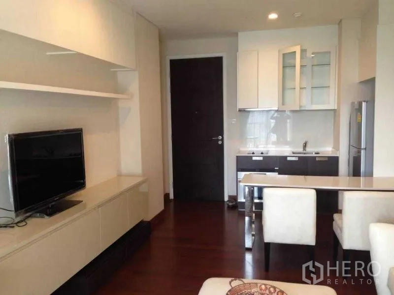 Condominium for rent or sale in Watthana, Bangkok - Cozy living room with TV console and shelving opposite a compact kitchenette and dining stools near the entry door.