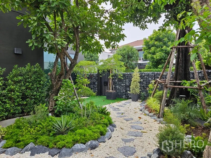 Detached House for sale in Thawi Watthana, Bangkok - Lush garden view with pebble path, stone fence and mature trees.