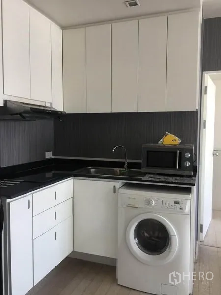 Condominium for rent in Watthana, Bangkok - Close-up of L-shaped kitchen with sink, cooktop hood, overhead storage, microwave, and in-unit washer.