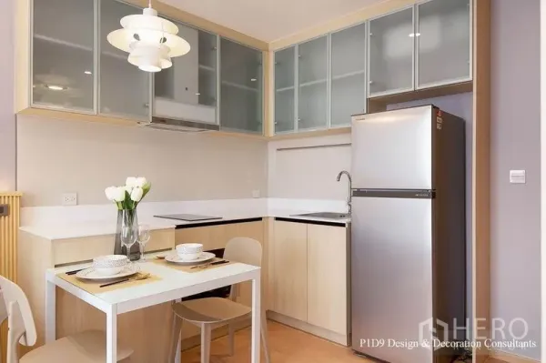 Condominium for rent in Watthana, Bangkok - Compact modern kitchen with glass‑front cabinets, white countertops, dining table for two and stainless fridge.