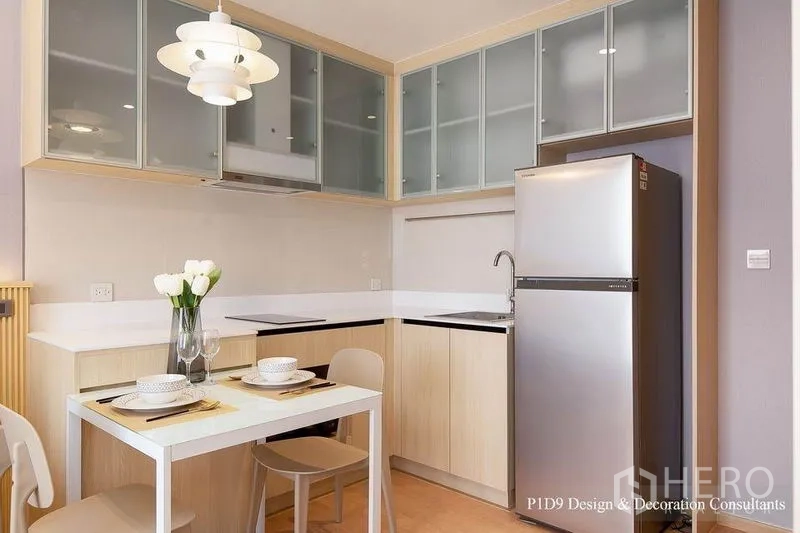 Condominium for rent in Watthana, Bangkok - Compact modern kitchen with glass‑front cabinets, white countertops, dining table for two and stainless fridge.