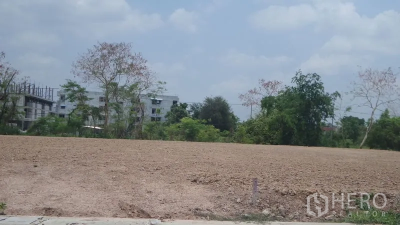 Land for rent in Lam Luk Ka, Pathum Thani - Prepared earth-fill land with neighboring low-rise buildings and mature trees.