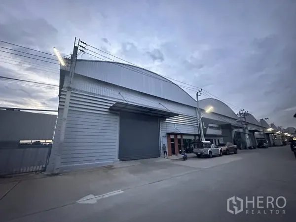 货栈 出租在Bang Phli, Samut Prakan - 傍晚的仓库立面，卷闸门关闭并有路灯照明。