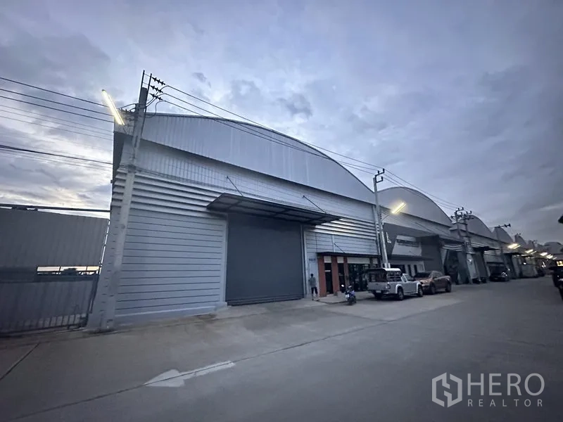 货栈 出租在Bang Phli, Samut Prakan - 傍晚的仓库立面，卷闸门关闭并有路灯照明。