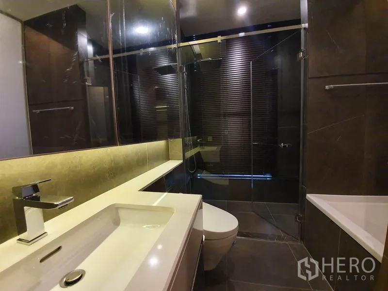 Condominium for rent in Huai Khwang, Bangkok - Contemporary bathroom with large mirror, vanity, glass-enclosed shower, bathtub, and dark tiles.