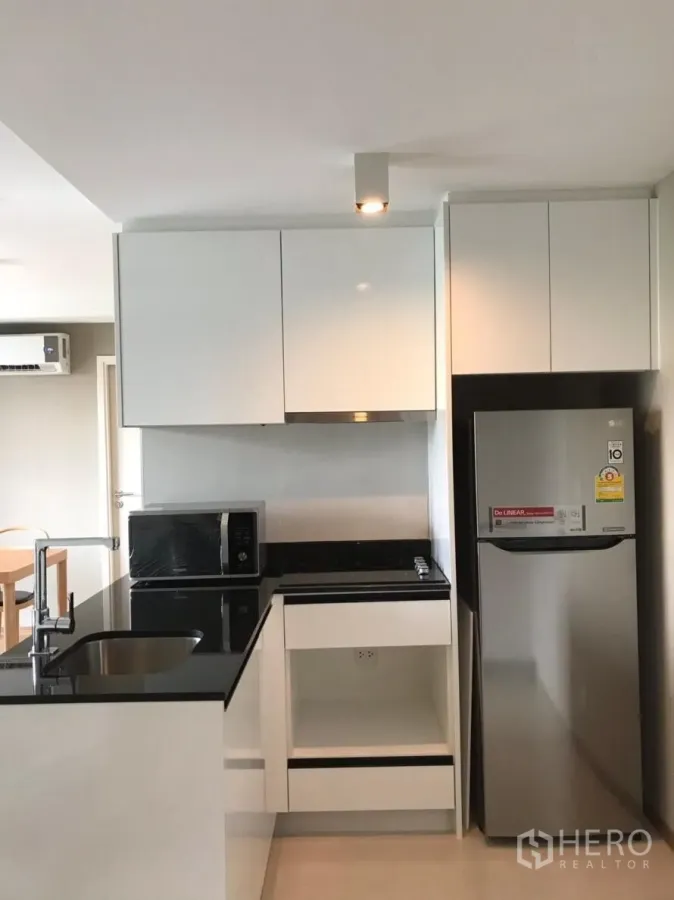 Condominium for rent in Watthana, Bangkok - Modern white kitchen with black stone countertop, sink, cooktop, microwave and stainless fridge.