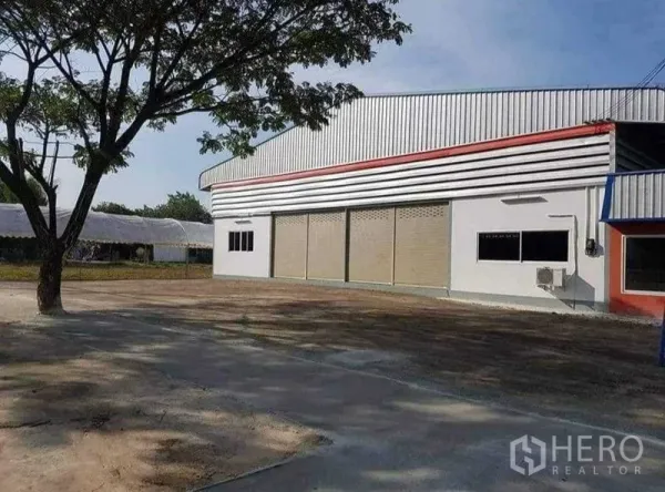 货栈 出租在Chon Buri - 萨塔希普金属板外墙仓库外观，多个卷闸门，宽阔混凝土场地与遮荫大树。