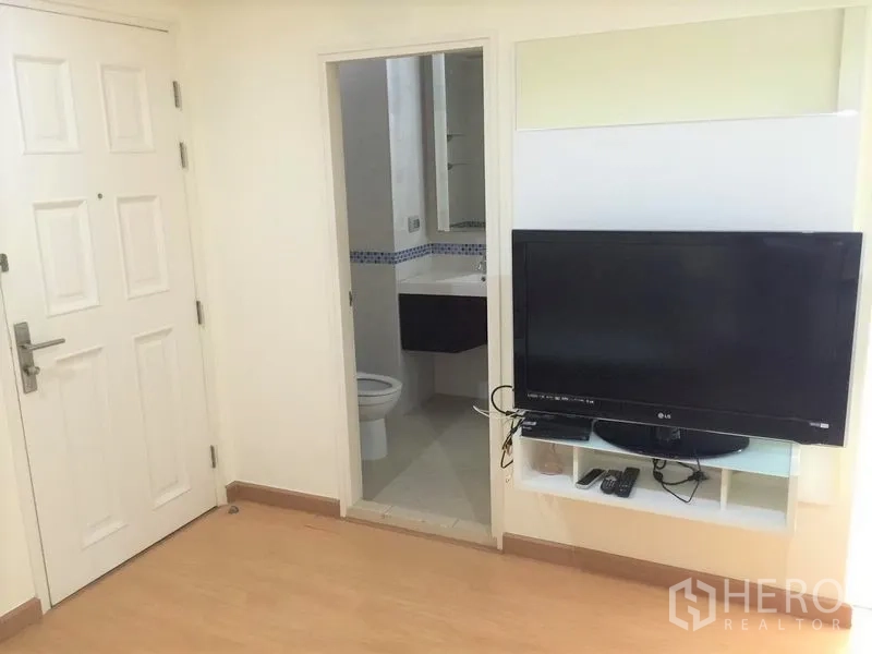 Condominium for rent in Watthana, Bangkok - Living area with wall-mounted TV console, light wood floors and a doorway leading to a tiled bathroom.