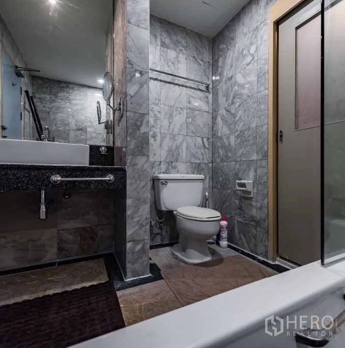 Condominium for rent or sale in Pathum Wan, Bangkok - Marble-tiled bathroom with vanity, toilet, and a bathtub with glass screen.