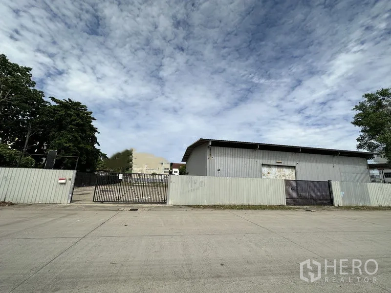 Warehouse for rent in Min Buri, Bangkok - Street view of a gated industrial compound with a metal fence and grey warehouse in Min Buri.
