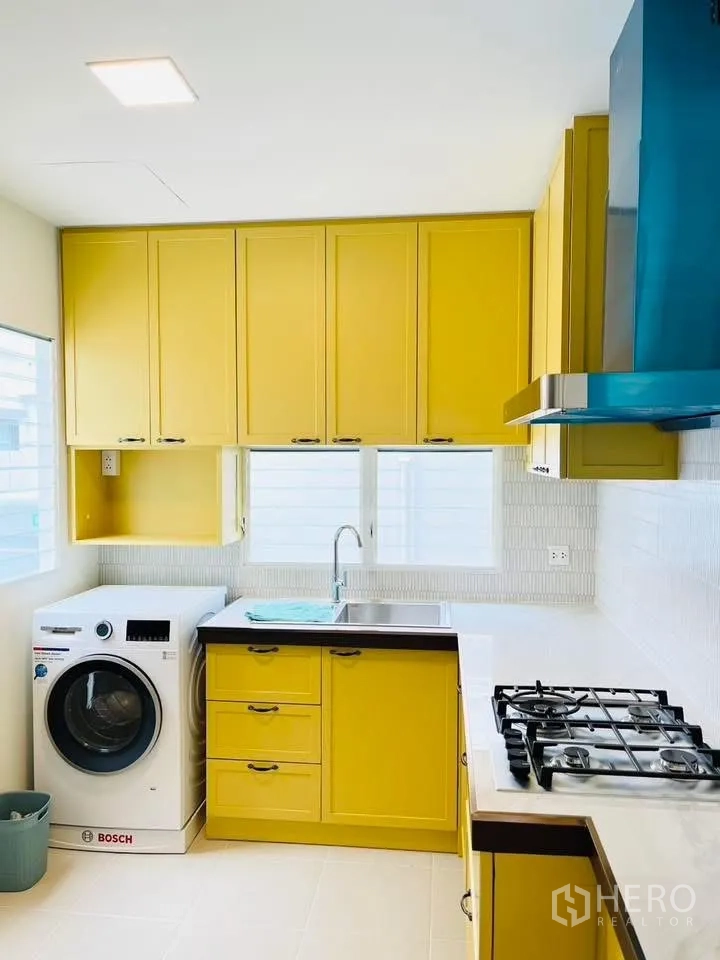 Detached House for rent in Bang Phli, Samut Prakan - L-shaped kitchen with yellow cabinets, washing machine, sink, and gas hob.