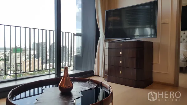 Condominium for rent in Watthana, Bangkok - Living area with floor-to-ceiling windows opening to a balcony and skyline view, TV on dark dresser and round coffee table.
