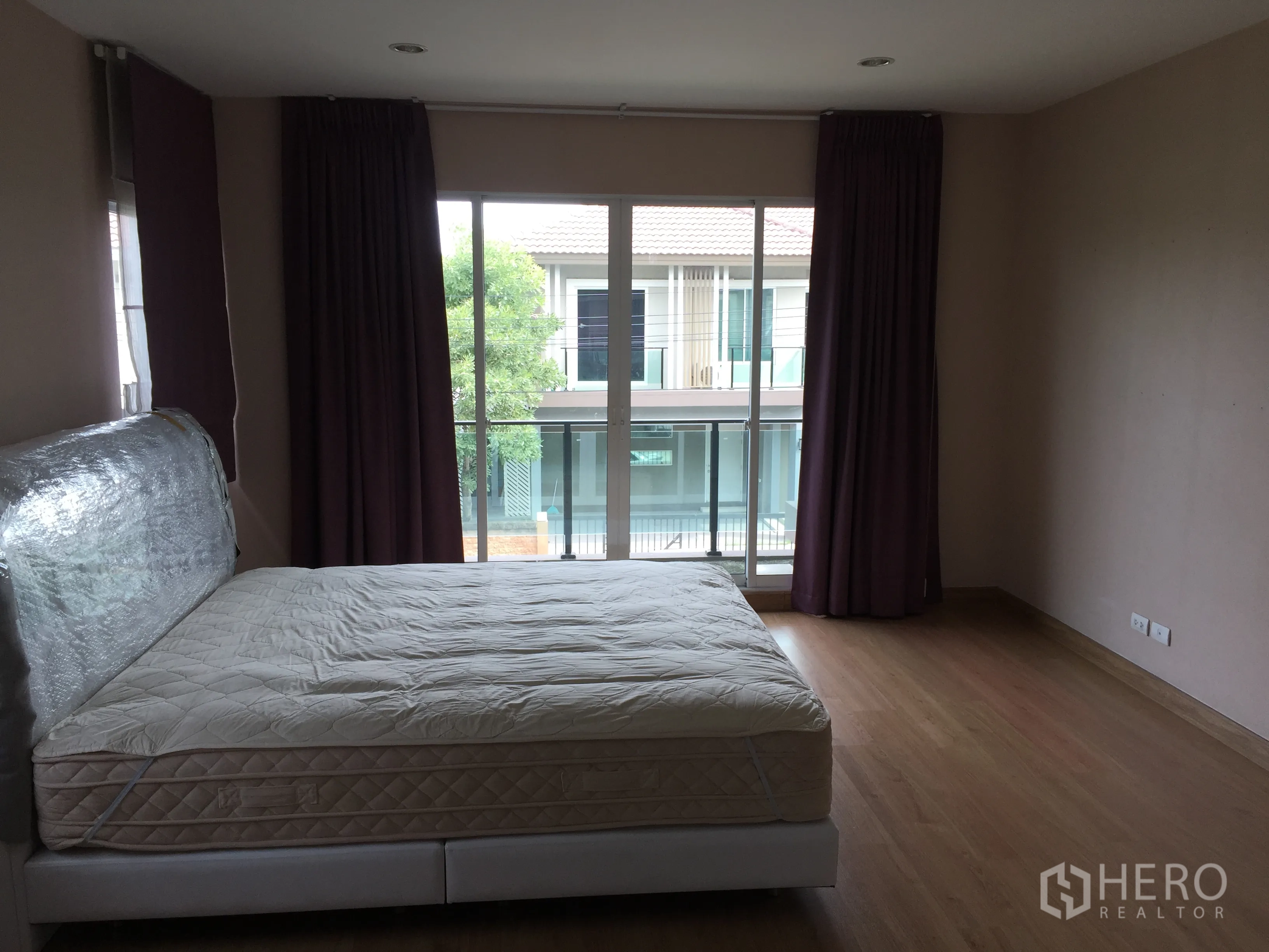 Detached House for rent in Pathum Thani - Spacious master bedroom with wood floors and balcony doors dressed with curtains.