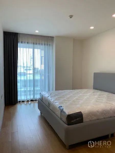 Condominium for sale in Khlong Toei, Bangkok - Cozy bedroom with large window, sheer curtains and light wood flooring.