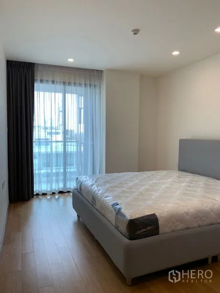 Condominium for sale in Khlong Toei, Bangkok - Cozy bedroom with large window, sheer curtains and light wood flooring.