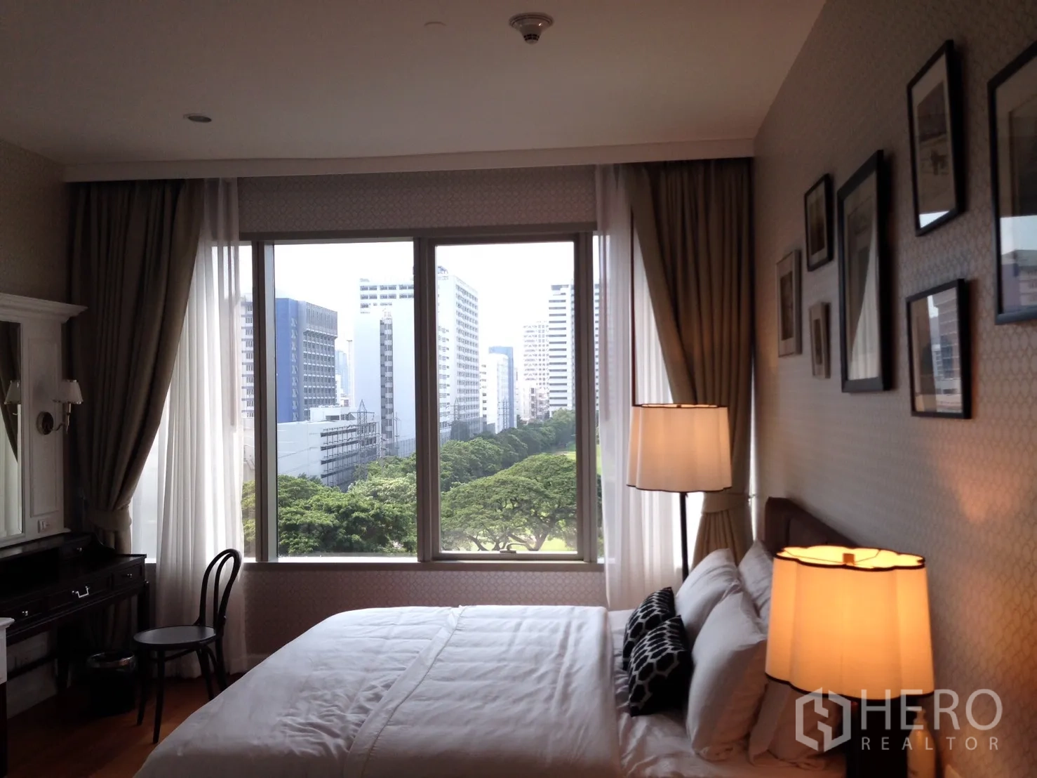 คอนโด ให้เช่าหรือขายในPathum Wan, Bangkok - ห้องนอนหน้าต่างบานใหญ่และโคมไฟหัวเตียง มองเห็นวิวสวนสีเขียวและสกายไลน์เมือง.