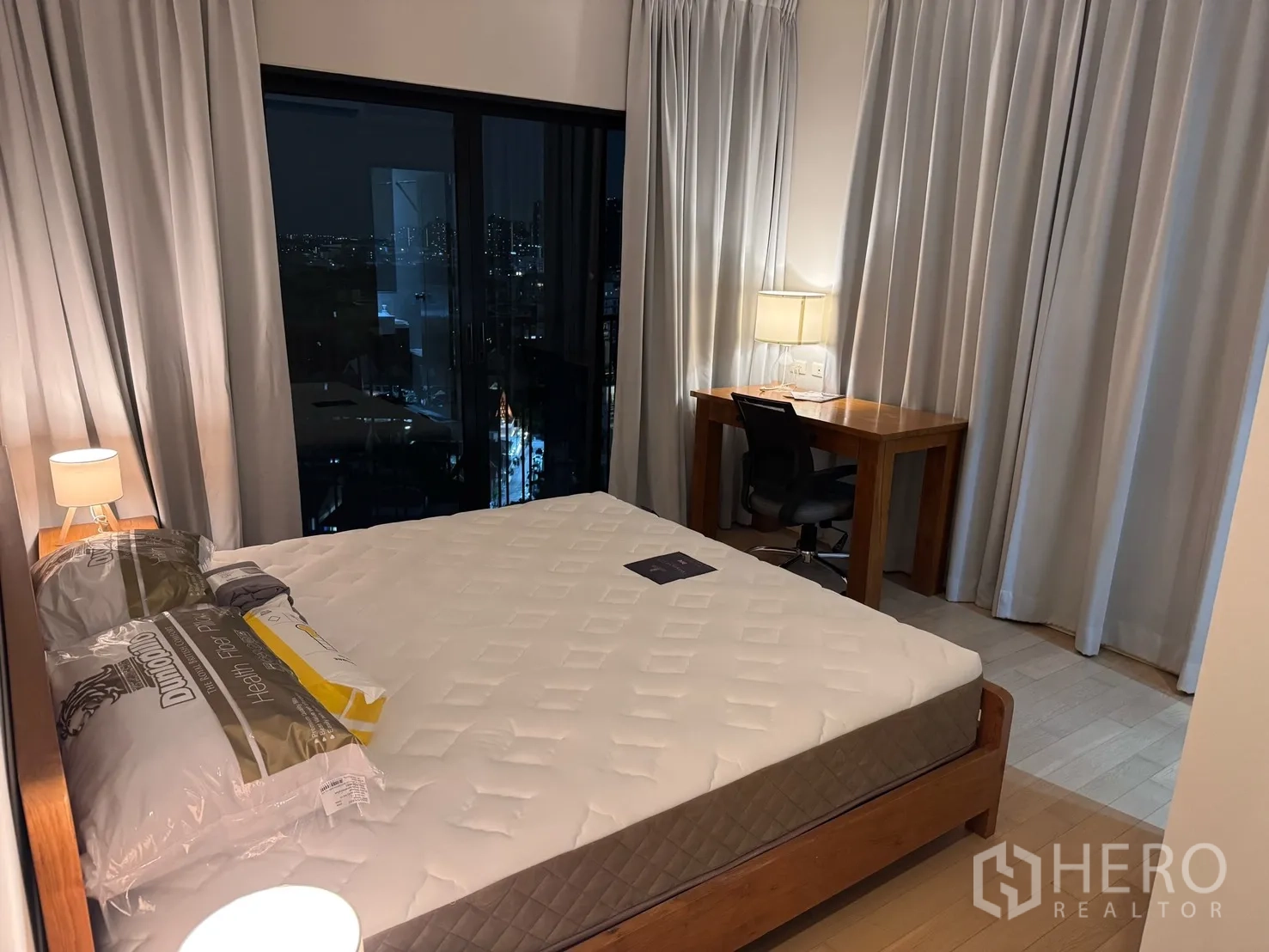 คอนโด for rent or sale in Watthana, Bangkok - ห้องนอนหลักเตียงขนาดใหญ่ โคมไฟหัวเตียง โต๊ะทำงานริมหน้าต่าง และวิวเมืองผ่านกระจกบานเลื่อน