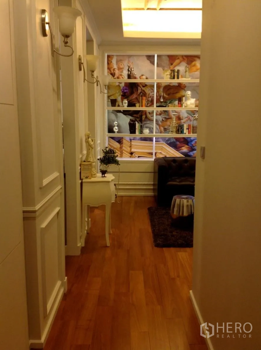 Condominium for rent in Watthana, Bangkok - Hallway with warm wood flooring leading to a living area featuring a backlit ceiling and art-filled display shelves.