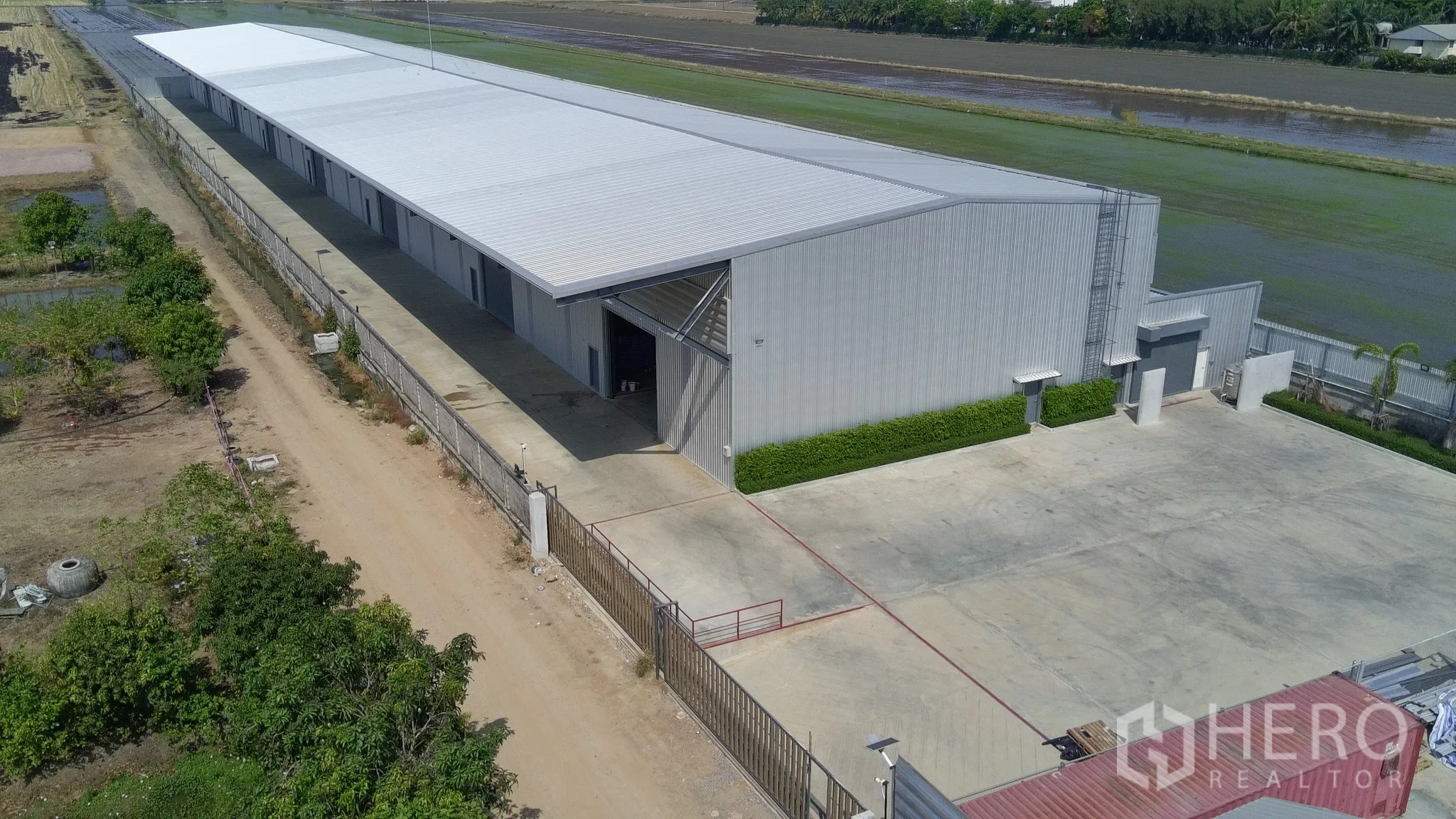Warehouse for rent in Lam Luk Ka, Pathum Thani - Aerial view of a long metal-clad warehouse with a gated entrance and wide concrete yard.