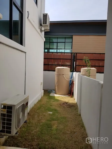 Detached House for sale in Samut Prakan - Side yard with grass strip, water tanks, AC condenser and privacy wall.