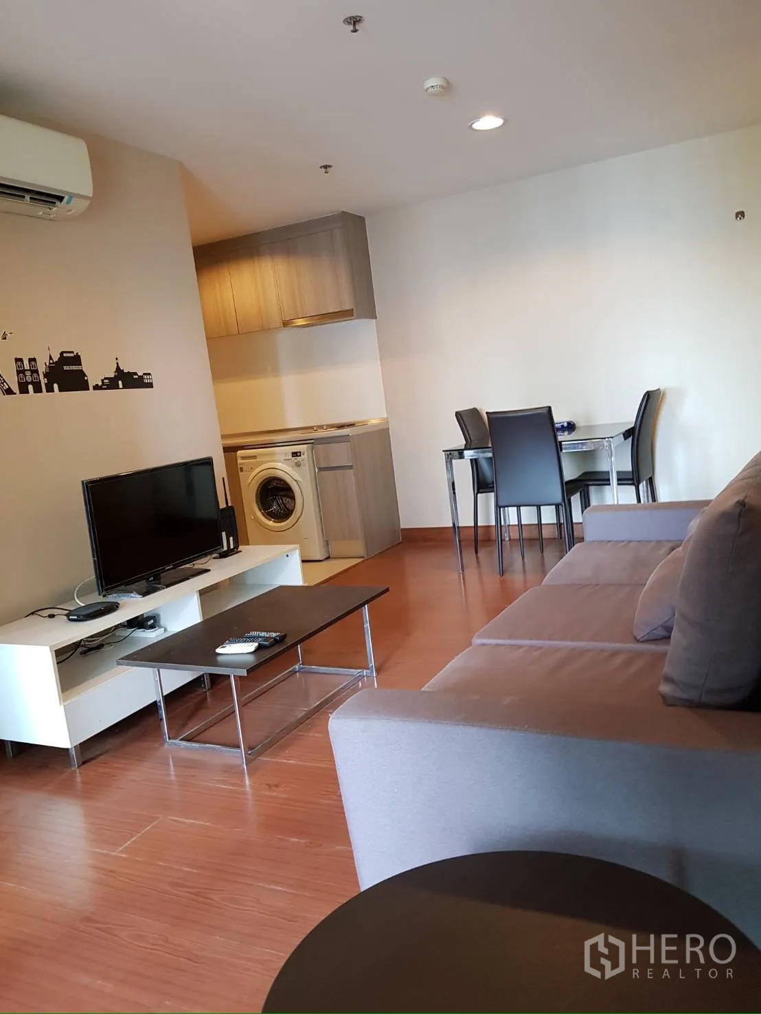 Condominium for rent in Huai Khwang, Bangkok - Living area with sofa, TV console, coffee table, and a washing machine by the kitchen.