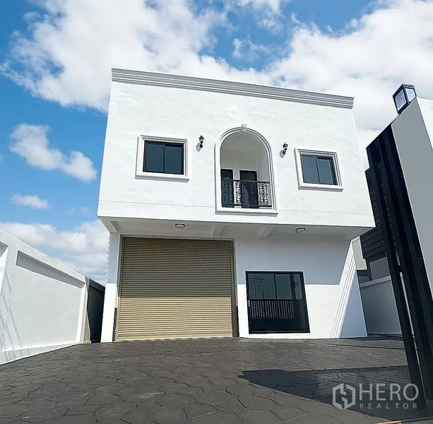 货栈 出租在Mueang Pathum Thani, Pathum Thani - 两层白色仓库外立面，拱形阳台、卷闸门与带门禁的车道，蓝天下。